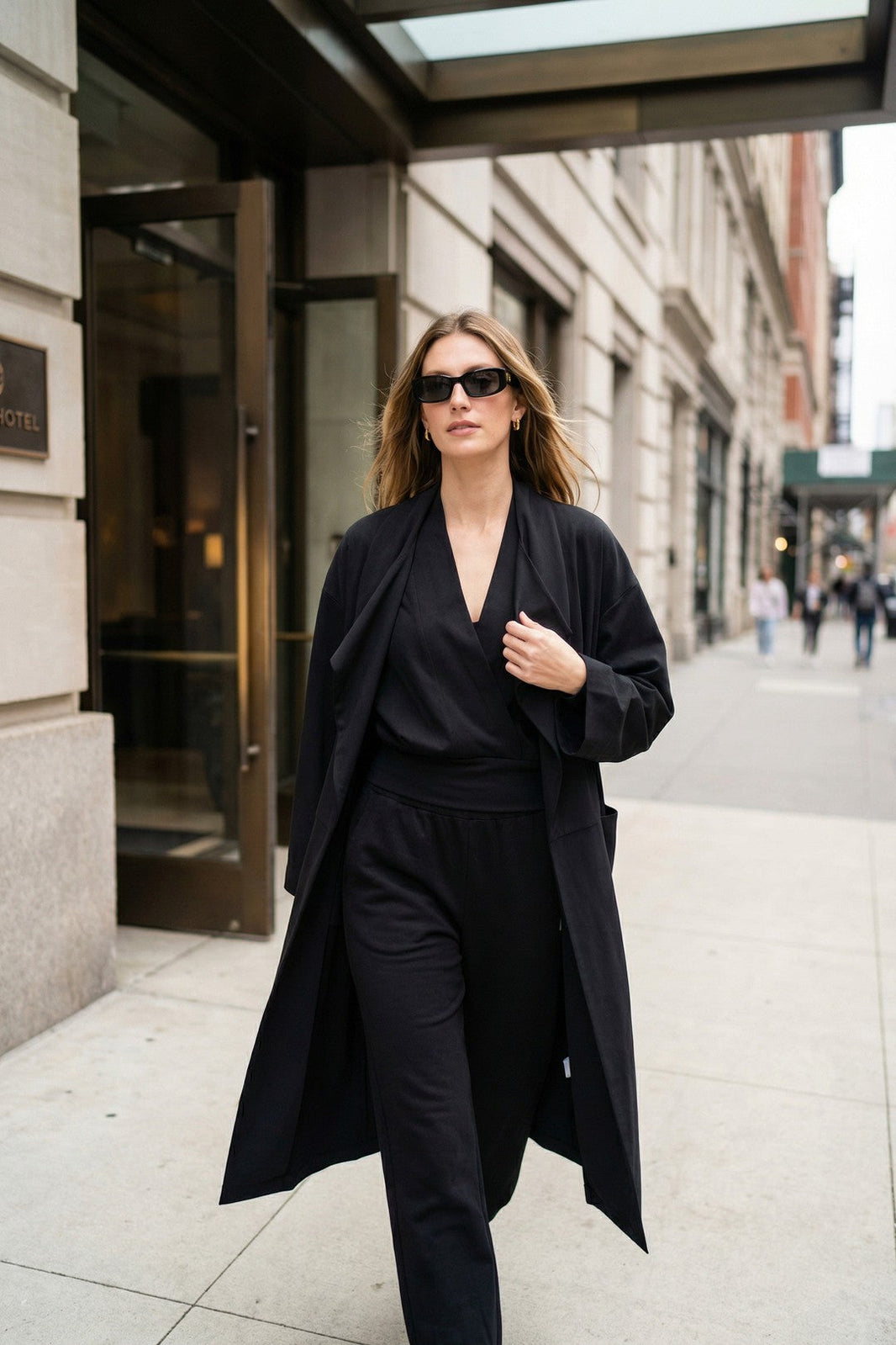 Woman walking on a city sidewalk wearing a long tailored coat over a coordinated outfit and sunglasses