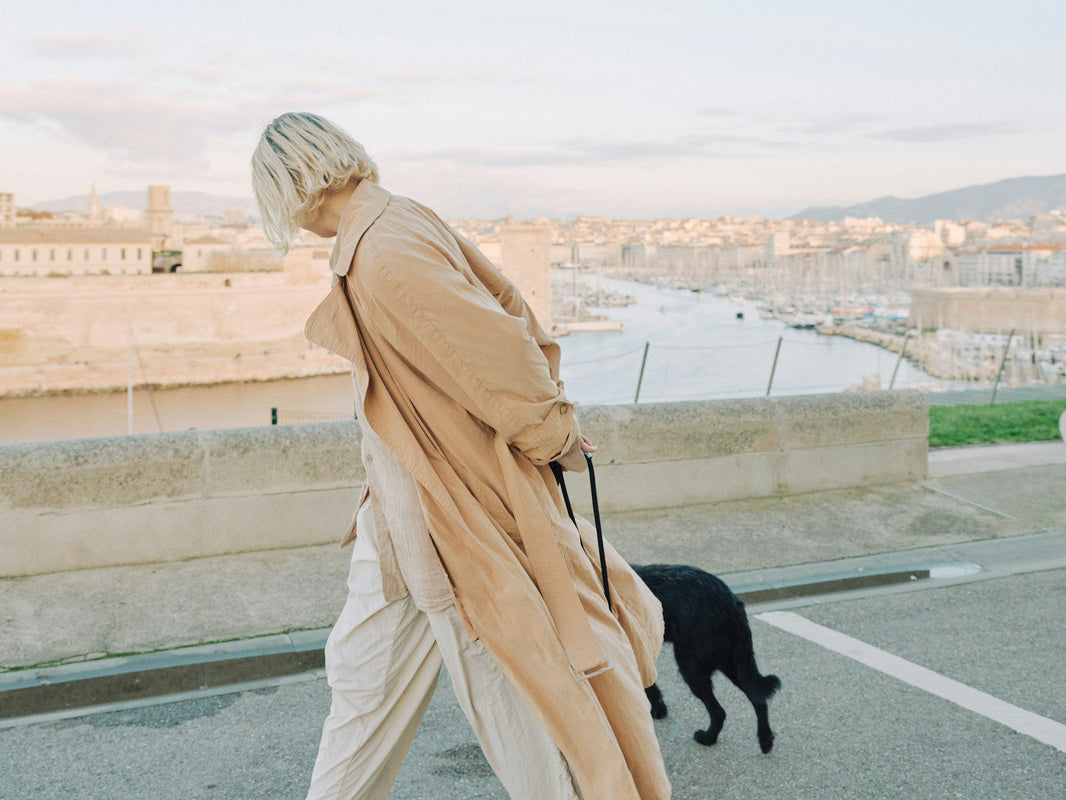 Woman walking along a waterfront promenade wearing a long lightweight jacket, with a cityscape in the background